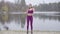 Wide shot of sportive brunette woman in purple sportswear and headphones listening to music and dancing on lake shore in
