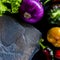 A wild multi-colored stone with a place for text lies on a black table next to fresh blue onions, yellow peppers, chili and