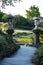 Winding garden path with Chinese style lanterns on pillars leading to water lilies floating on pond with trees in background