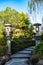 Winding garden stone pathway with Chinese pillars, weeping willow tree, shrubs against blue sky