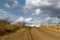 A winding wide road with turns of dust, earth and sand, among dry vegetation and grass. On the background of a beautiful cloudy sk