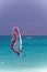 Windsurfer man on board with red colored sail on ocean on sunny day on blue sky background. Recreational water sports during idyll