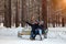 In the winter afternoon, a loving couple in jackets and hats are sitting on a bench in the woods in the snow hugging the guy shows