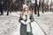 Winter fashion. Portrait of young woman wearing long green coat with scarf, hat, mittens in snowy park