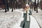 Winter fashion. Portrait of young woman wearing long green coat with scarf, hat, mittens in snowy park.