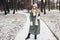 Winter fashion. Young woman wearing long green coat with scarf, hat, mittens and boots in snowy park