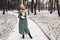 Winter fashion. Young woman wearing long green coat with scarf, hat, mittens and boots in snowy park.