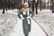 Winter female fashion. Young woman wearing long green coat with scarf, hat, mittens in snowy park.