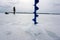 Winter fishing. Silhouette of a fisherman in the vastness of a frozen reservoir. In the foreground is the process of drilling a