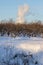 Winter landscape with an apple orchard and a column of smoke above it against a blue sky on a clear sunny frosty day.