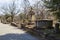 A winter landscape in the graveyard with headstones, graves, bare winter trees, lush green trees, plants and flowers