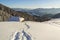 Winter landscape of mountain valley on frosty sunny day. Footprint path in white deep snow leading to small old wooden shepherd h