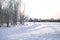 Winter landscape - white snow with traces of shoes and skis on the field. The ski track and road skirting the forest with bare tre