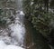 Winter scenery with snowfall and mountain stream running through picturesque snowy forest. River coursing in canyon through