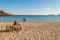 Winter sea beach and calm sea on a sunny day. A lonely elderly woman sits in a deck chair facing the sea. A loving couple stands