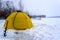 A winter yellow fishing tent stands next to an ice pack on snow and ice in a Blizzard.