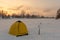 Winter yellow tent with ice screws near fisherman standing on the ice of a Siberian river on sunset.