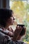 Wistful millennial woman with cup of tea and woolen plaid sitting near  window and looking at autumn landscape outside. Fall mood