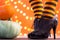 Witch legs in striped stockings and high heel shoes with pumpkins on an orange background, bokeh. Halloween. Copy space.