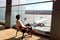 Woman in the airport lounge at a large window on a chair watches the runway. Aircraft of the RED WING company