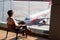 Woman in the airport lounge at a large window on a chair watches the runway. Aircraft of the RED WING company