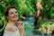 woman alone with elegant hair bun in white off shoulder blouse enjoys nature green park with river smiling and looking into camer