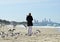Woman alone walking on beach surrounded by flock sea birds & distant city life