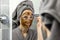 Woman applying mud facial mask with brush in front of mirror in bathroom at home. skin care