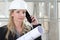 Woman architect or construction engineer talk on the mobile phone wear helmet and holds blueprint inside a building site with