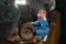 A woman auto mechanic sits near a disassembled car, dressed in overalls and safety glasses, holds a wheel and looks into