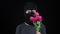 A Woman in a balaclava mask is standing with flowers. Bandit sniffs a bouquet of pink flowers on a black background.