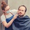 Woman barber in medical mask shaving a client in a home bathroom with a safety razor. Concept of unkempt appearance and problems