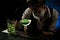 Woman bartender in mask and black gloves carefully add ingredient to smoky glass with green cocktail.