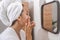 woman in bathrobe, skincare routine, applies moisturizer in front of the mirror, home self-care.