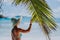 Woman on the beach touching palm tree leaf shadow wearing blue hat. Luxury paradise recreation vacation concept