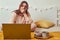 A woman on a bed falls asleep in the evening room with a laptop. The girl works on the computer and has dinner on the bed