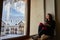 Woman being cold sits on windowsill, warming up with tea mug, looking at snow covered mountains. Female drinks coffee in