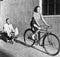 Woman on a bicycle pulling a grown man on a toy tricycle