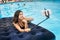 Woman in bikini smiling and taking selfie photo on the phone with selfie stick on a mattress in the pool at the resort.