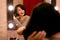 A woman with black hair straightens her hair at a dressing table with a mirror