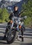 A woman in a black leather biker jacket with a carbine rifle on a chopper motorcycle in Greece on a road in the forest in the moun