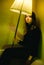 A woman in black with vibrant makeup sits alone at home holding a floor lamp in darkness. She appears sad and contemplative.