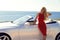 Woman with blond hair in red dress posing beside luxury auto