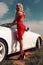 Woman with blond hair in red dress posing beside luxury auto