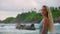 Woman in blue dress enjoys serene tropical beach, observes waves, lighthouse. Blonde woman strolls along shore, admires