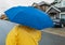 Woman with blue umbrella walking in the rain on a street. Unrecognizable person in yellow rain jacket with umbrella