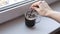 Woman brews instant coffee in a transparent glass cup on a window sill in the morning and stir it with a spoon