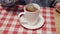 A woman brews a tea bag in a white cup on a plaid tablecloth.