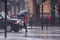 A woman in a brown coat under an umbrella crosses a busy street in heavy rain. Heavy precipitation in the city. Flooding