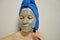 A woman with a brush applies a clay mask to her face with blue towel on her head